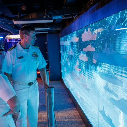 A person in a naval uniform observes a digital display wall with lit models of ships in a dark, futuristic room.