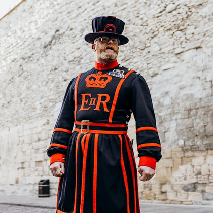 A man in traditional black and red uniform stands in front of a stone wall. His attire features a crown symbol and "ER" lettering.