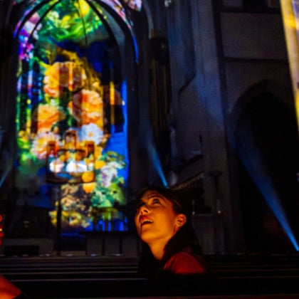 A person looks up in a church with colorful light projections on the walls and stained glass windows.