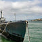A docked submarine tied with a rope, with calm water and a distant coastline under a cloudy sky.