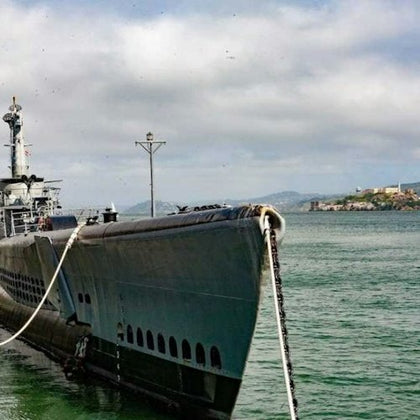 A docked submarine tied with a rope, with calm water and a distant coastline under a cloudy sky.