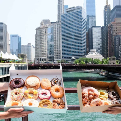 Two hands holding open boxes of assorted donuts with a city skyline and river in the background.
