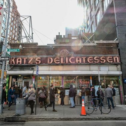 People lined up outside Katz's Delicatessen on a busy street with bicycles and a construction cone near the entrance.