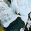 Frozen waterfall cascading into a partially frozen river surrounded by snow and ice-covered cliffs.