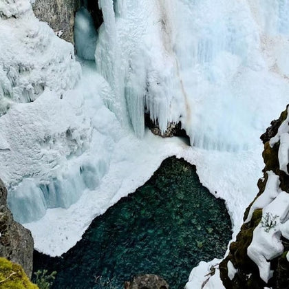 Frozen waterfall cascading into a partially frozen river surrounded by snow and ice-covered cliffs.