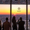 Four people stand inside a high-rise building, watching the sunset over a cityscape through large windows marked "West."