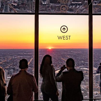 Four people stand inside a high-rise building, watching the sunset over a cityscape through large windows marked "West."