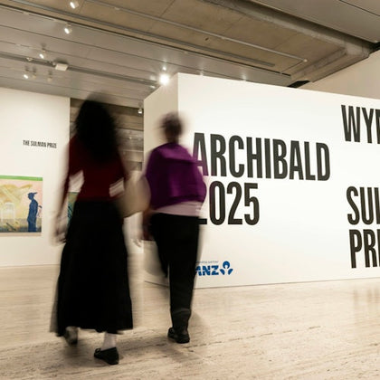 Two people walking in a modern art gallery with visible signage for the Archibald 2025 and Wynne prizes.
