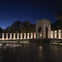 Night view of a war memorial with illuminated columns, a central archway, and a fountain in the foreground.