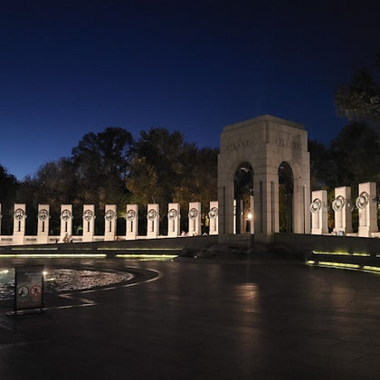 Night view of a war memorial with illuminated columns, a central archway, and a fountain in the foreground.