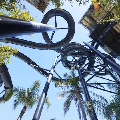 A view from below of a complex water slide structure with black tubes, surrounded by palm trees under a clear blue sky.