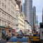 A busy urban street with yellow taxis, pedestrians, and historic buildings alongside modern skyscrapers under a cloudy sky.