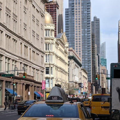 A busy urban street with yellow taxis, pedestrians, and historic buildings alongside modern skyscrapers under a cloudy sky.