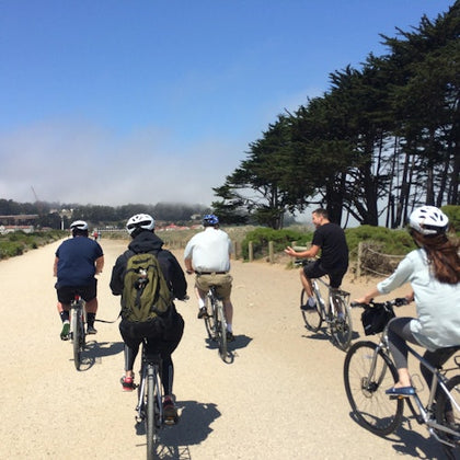 A group of people riding bicycles on a gravel path surrounded by greenery and trees under a sunny, partly cloudy sky.