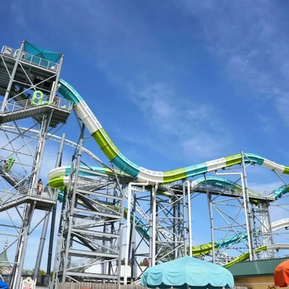 A tall water slide with green and blue sections supported by a metal structure under a clear blue sky.
