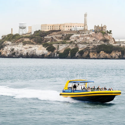 A group of people seated on a boat, raising their hands and smiling, with water and a cloudy sky in the background.