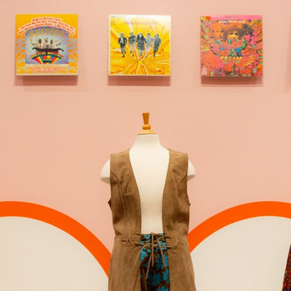 A mannequin dressed in a tan vest and blue patterned pants is displayed in front of a pink wall with album covers above.