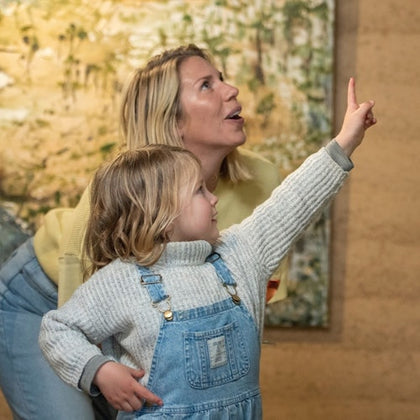 A woman and a young child point and look up while indoors, with a blurred painting in the background.
