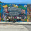 A group of people posing in front of a colorful mural with the text "Wynwood Mural" and various vibrant illustrations.