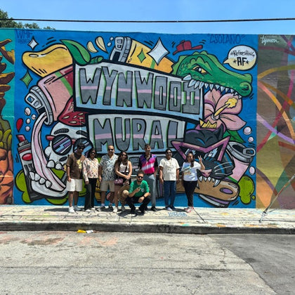 A group of people posing in front of a colorful mural with the text "Wynwood Mural" and various vibrant illustrations.