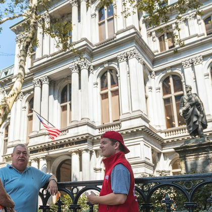 Two men stand in front of a historic building with a statue and an American flag, engaging in conversation on a sunny day.
