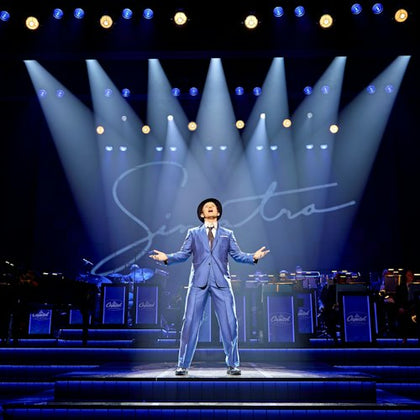 A performer in a blue suit and hat sings passionately on a stage under spotlights, with a band and "Sinatra" script behind.