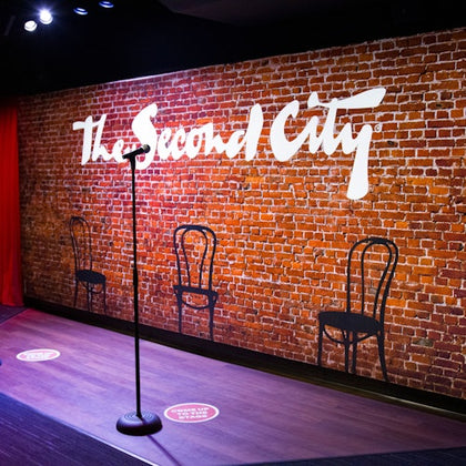 A brick wall with "The Second City" in white text, two chairs, and a microphone stand on a stage with social distancing markers.