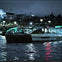 A boat on a river at night with illuminated city buildings and a bridge in the background.