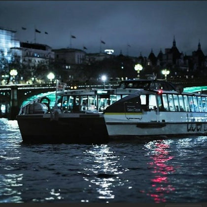 A boat on a river at night with illuminated city buildings and a bridge in the background.