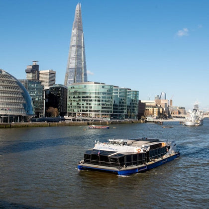 A boat on a river with modern buildings and the Shard skyscraper in the background, under a clear blue sky.