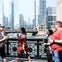 People walking on a bridge over a river with skyscrapers and a tour boat in the background on a sunny day.