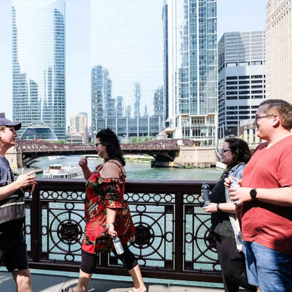 People walking on a bridge over a river with skyscrapers and a tour boat in the background on a sunny day.