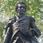 A bronze statue of a man in historical attire, surrounded by green leaves and trees.