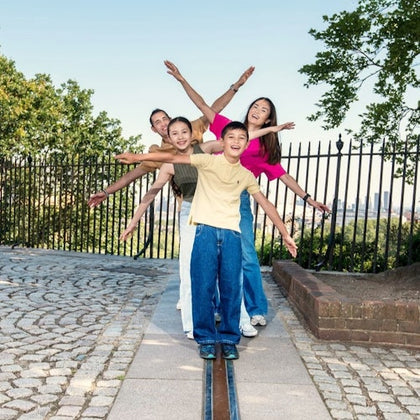 Four people stand in a line with their arms outstretched, smiling, and posing by a fenced area in a park with cobblestone paths and trees.