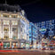 A busy urban street at night with festive Christmas lights, a large building, and double-decker buses in motion.