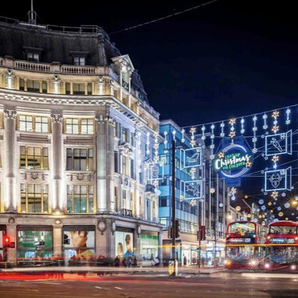 A busy urban street at night with festive Christmas lights, a large building, and double-decker buses in motion.