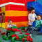 Children playing with large, colorful interlocking blocks near a playground slide. An adult supervises while another child enters a play area.