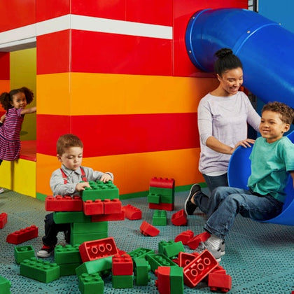 Children playing with large, colorful interlocking blocks near a playground slide. An adult supervises while another child enters a play area.