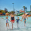 A family of four walks through shallow water at a water park, with colorful slides and attractions visible in the background.