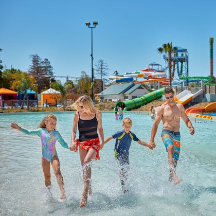 A family of four walks through shallow water at a water park, with colorful slides and attractions visible in the background.