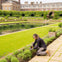 A person gardening near a manicured lawn and reflecting pool in a formal garden, with a large building in the background.