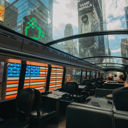 Inside a bus with a glass roof, showing a view of city buildings, a billboard, and an American flag pattern on the bus windows.