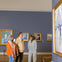 Three people observe paintings in an art gallery, with framed artwork on blue walls and a large painting to the right.