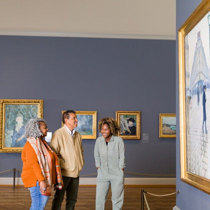 Three people observe paintings in an art gallery, with framed artwork on blue walls and a large painting to the right.