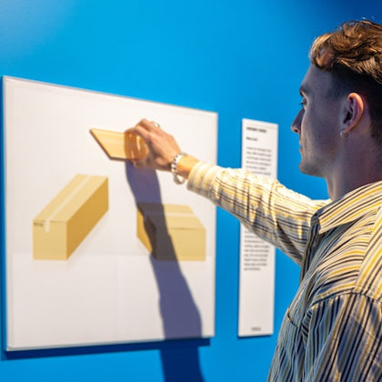 A person interacts with a tactile exhibit on a blue wall, touching a display featuring raised yellow shapes.