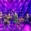 Performers on stage in vibrant costumes singing under purple and yellow lighting, with a lively backdrop and set.