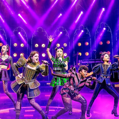 Performers on stage in vibrant costumes singing under purple and yellow lighting, with a lively backdrop and set.