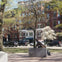 A bronze statue of a man seated on a stone pedestal in a park with trees and a red brick building in the background.