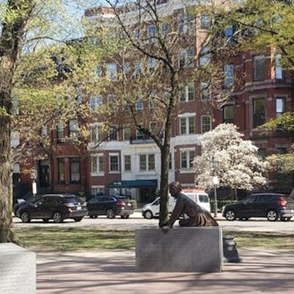 A bronze statue of a man seated on a stone pedestal in a park with trees and a red brick building in the background.