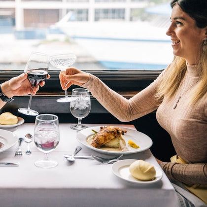 Two people seated at a restaurant table by a window, toasting drinks over a meal, smile and engage in conversation.